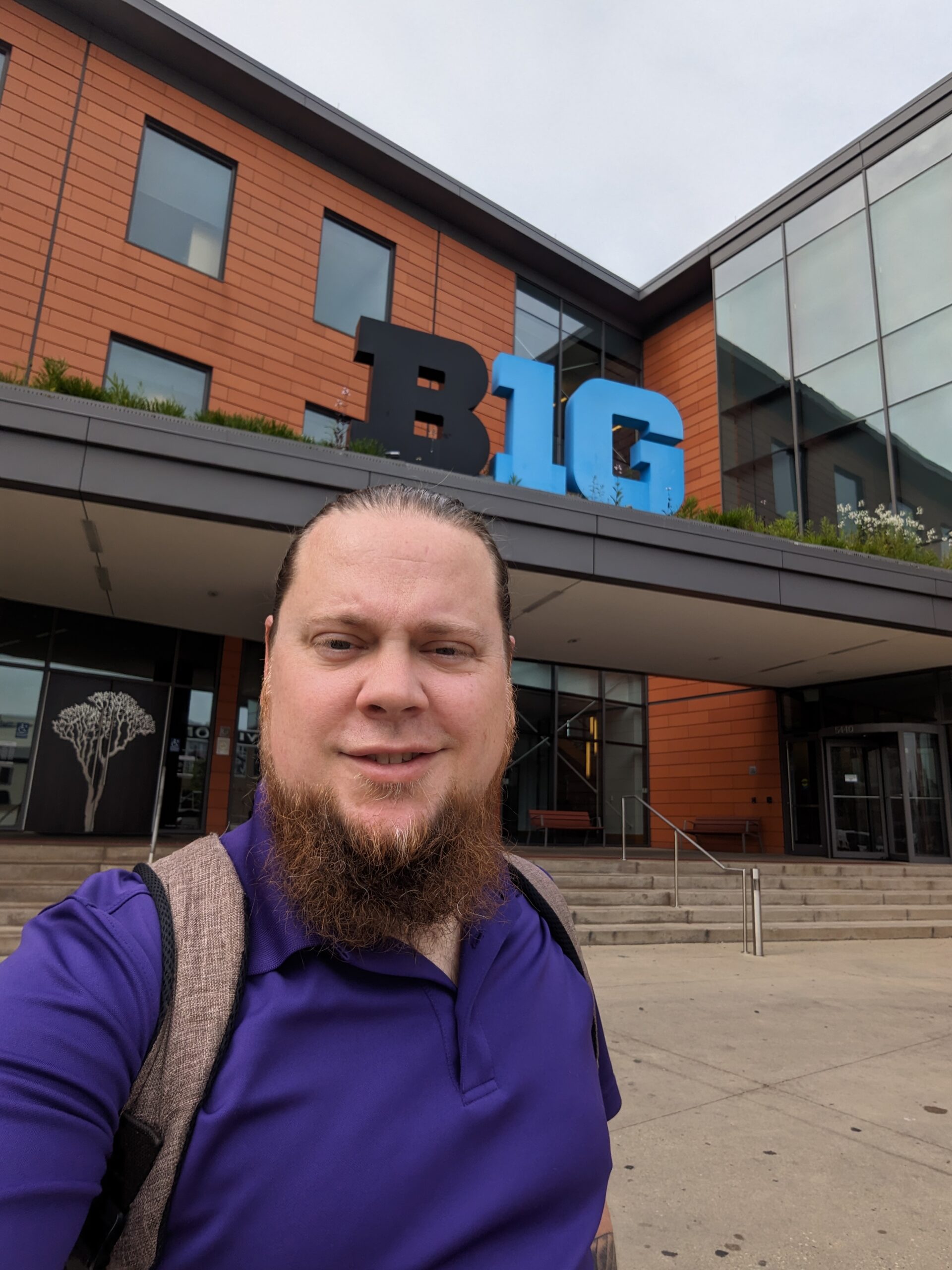 Evan Bradley standing in front of the Big Ten conference center in Rosemont, IL. He is wearing a purple shirt and standing in front of a building displaying the B1G logo.